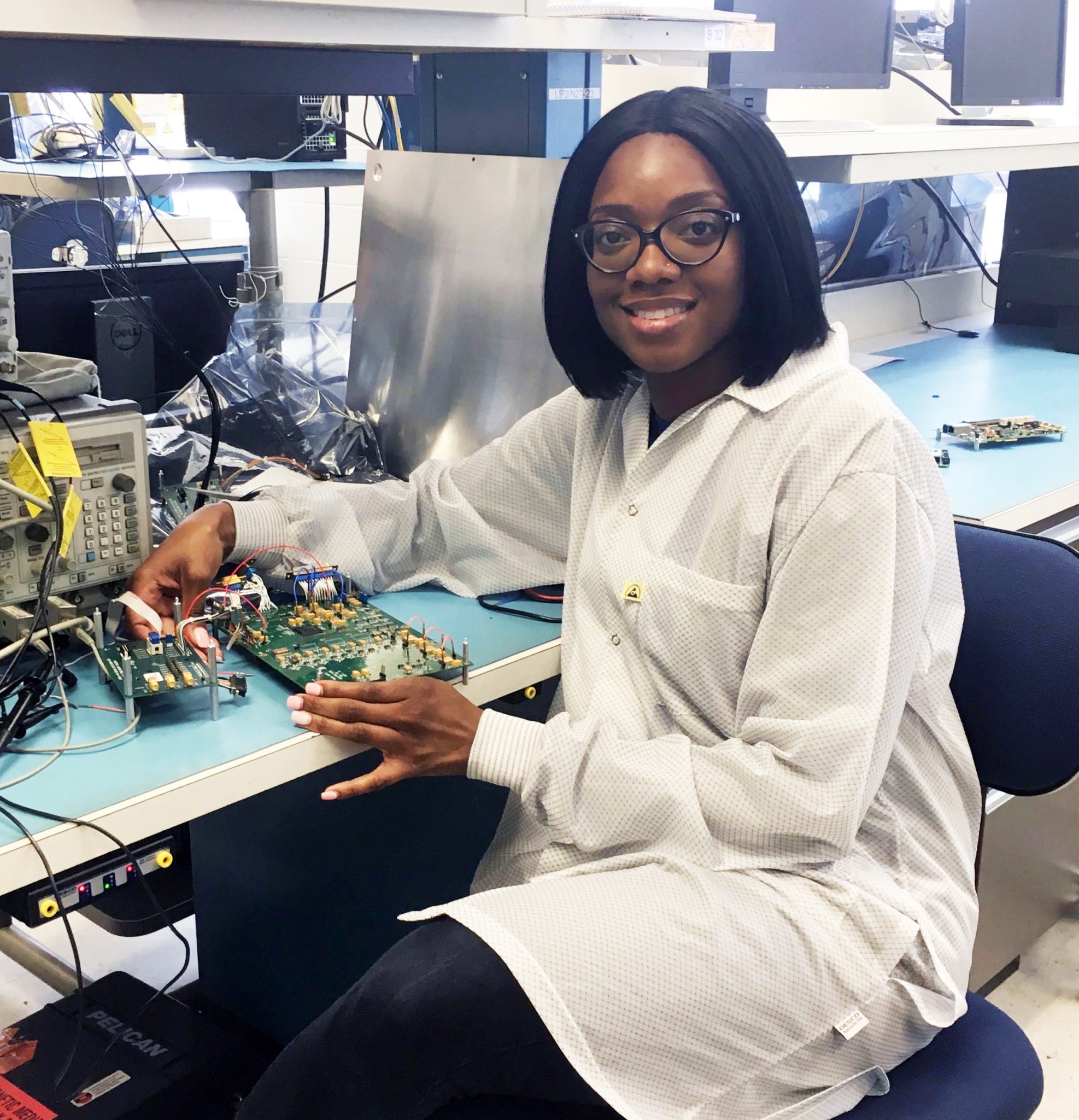 Sanetra is seated in front of a desk topped with some hardware she's been working on. She is smiling at the camera.