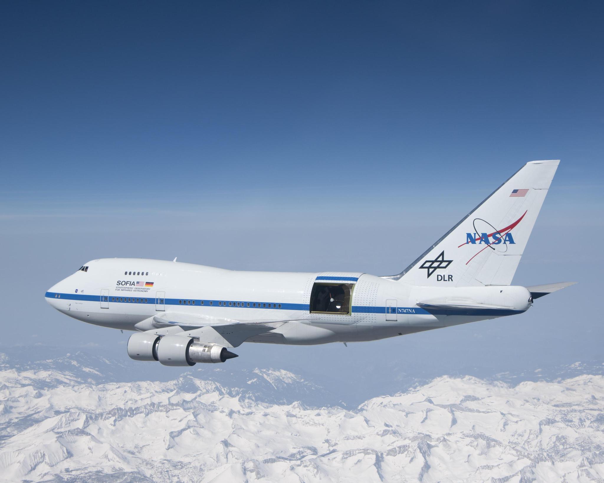 A white airplane floats over snowy mountains. The airplane has NASA insignia and a large opening on its side, where a telescope tool looks outward. 