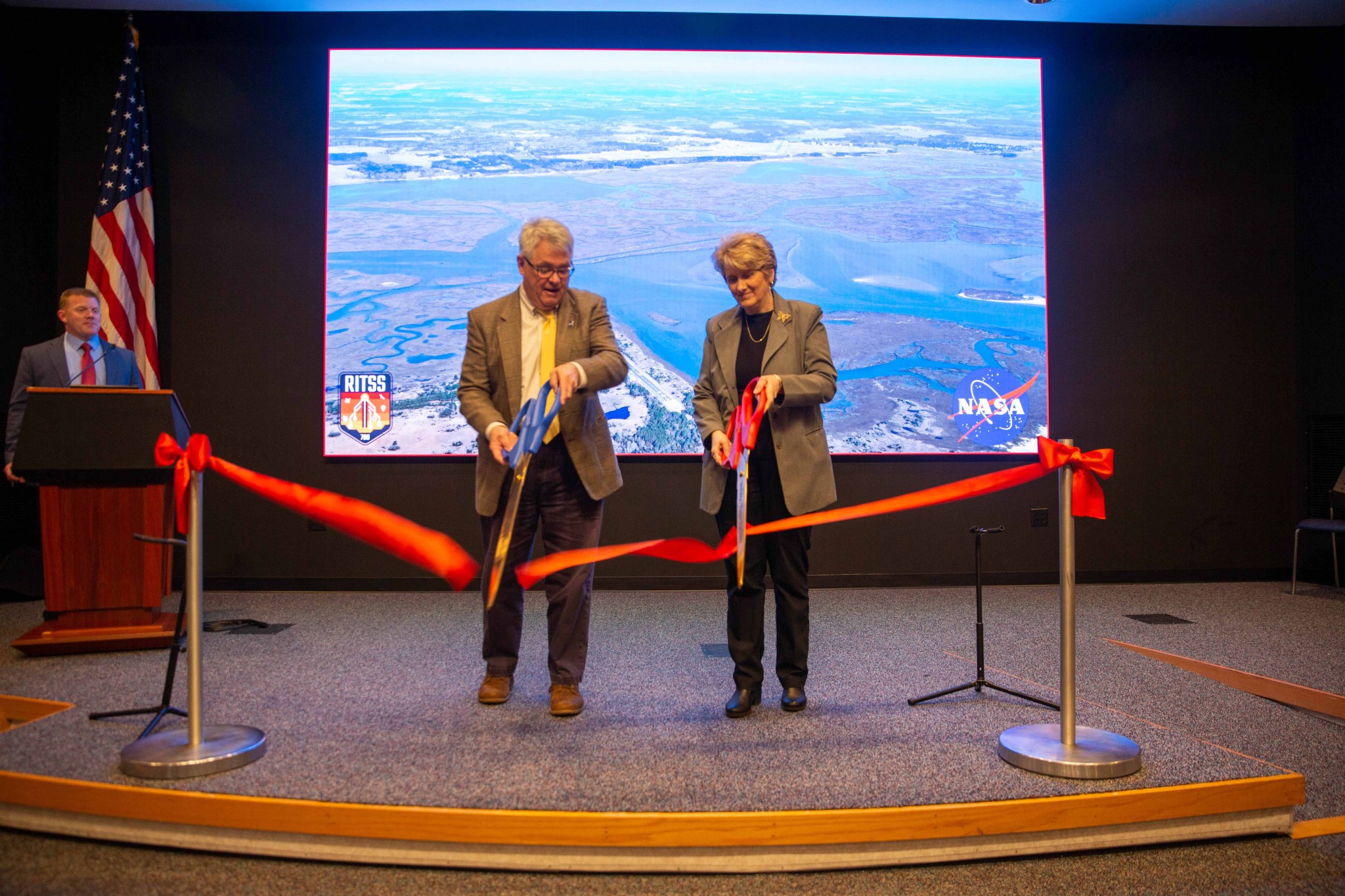 A white man and white woman each use a large pair of scissors to cut a red ribbon. They are standing on a stage in front of a large screen with an aerial image of a marsh area. Another white man stands behind them to the left with an American flag.