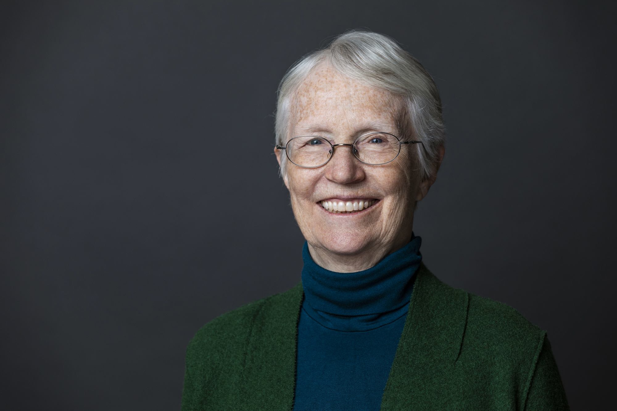 A headshot of Cynthia Rosenzweig. She is smiling, has short gray hair and glasses, and is wearing a navy blue turtleneck and green blazer.