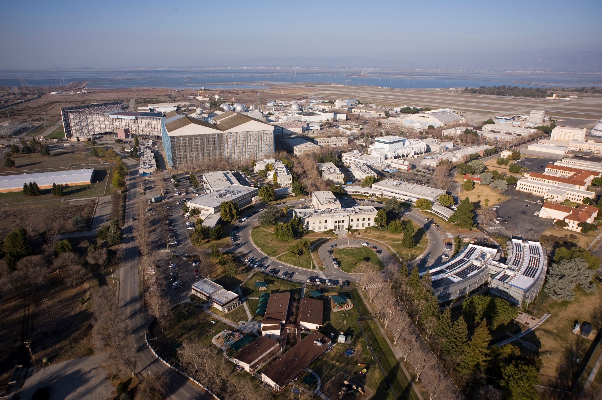 A view from above of a group of buildings around a circle. In the background there's larger buildings. At the horizon, there's water with mountains in the background.