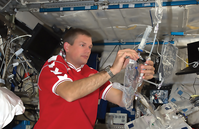 An astronaut holding a plastic bag connected to tubing in the interior of the International Space Station.
