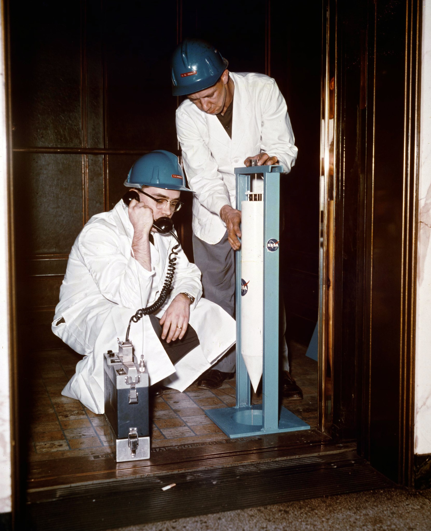 Two men dressed in lab coats, one kneeling and the other standing with large orange metal tubes