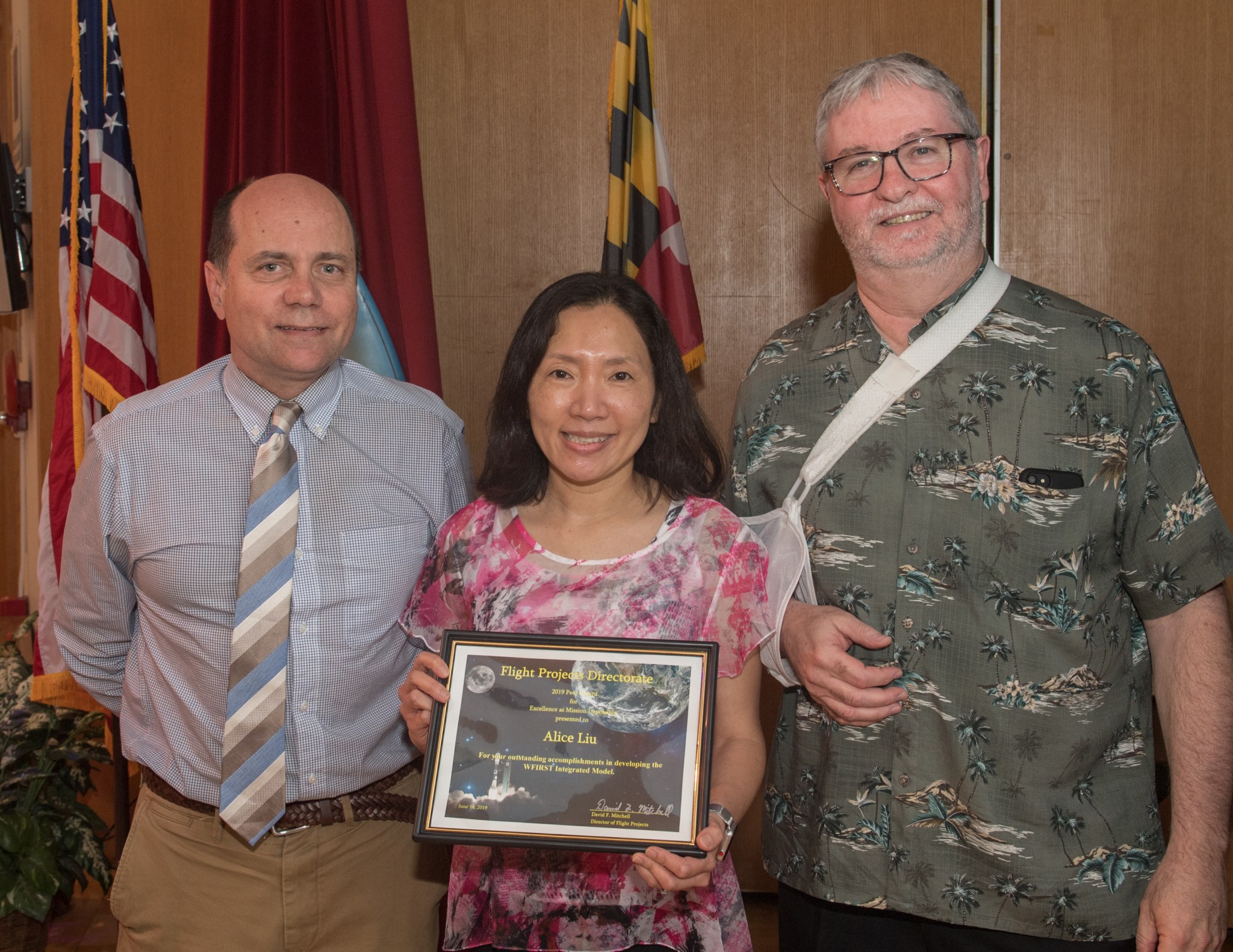 An Asian woman wearing a pink blouse holds a plaque between two white men. The man on the left wears a blue shirt and striped tie. The man on the right has gray hair/beard, and wears a green Hawaiian shirt and has his right arm in a sling.