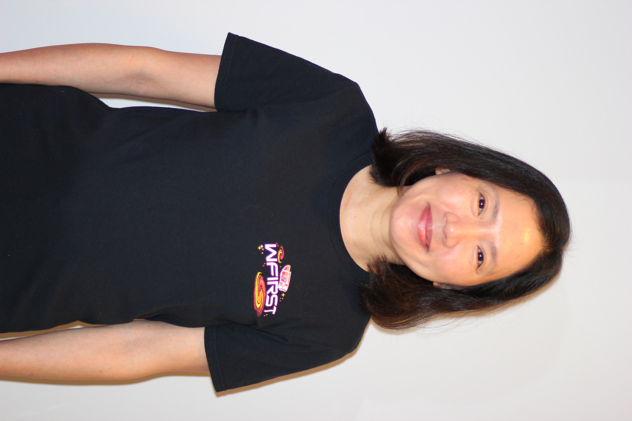 An Asian woman with shoulder-length dark hair stands in front of a white background, smiling. She is wearing a black T-shirt with the WFIRST logo on the left chest.