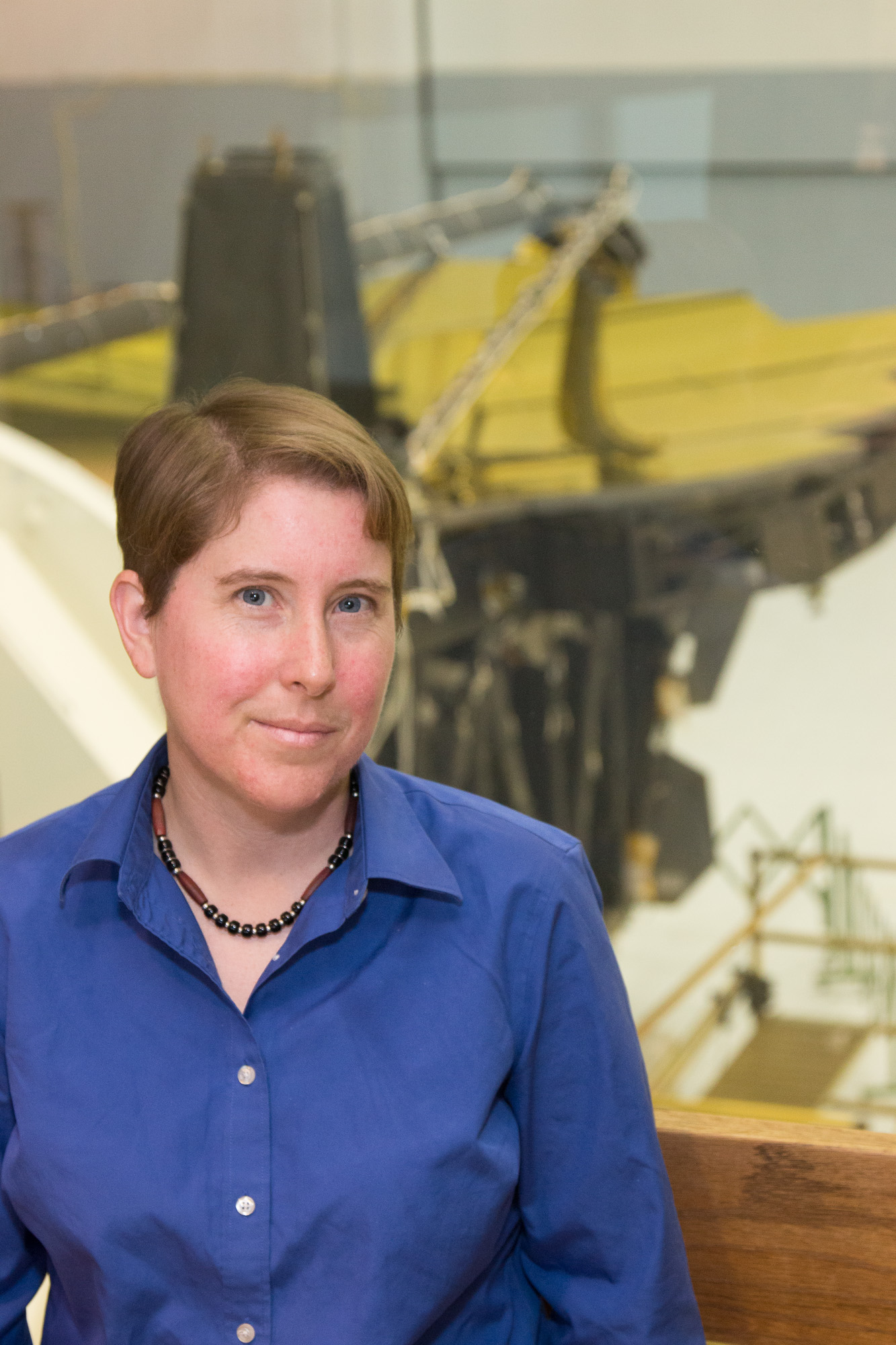 A white women with cropped short red hair and a blue button down shirt. They are standing on a balcony, and in the background in the golden mirror section of the James Webb Space Telescope laid down.