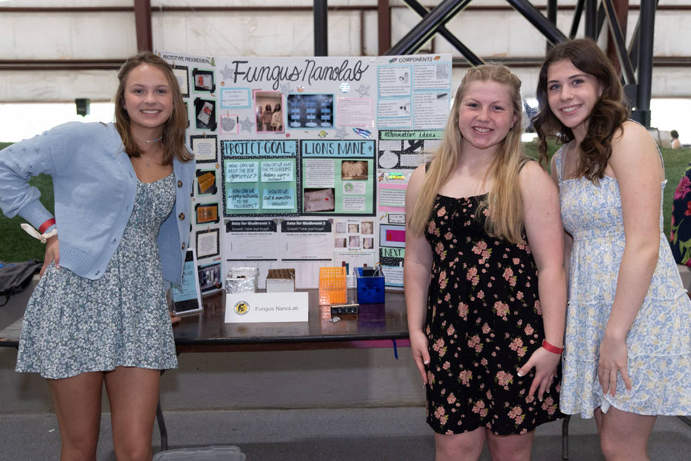 Three young women pose in front a poster mounted on a table.