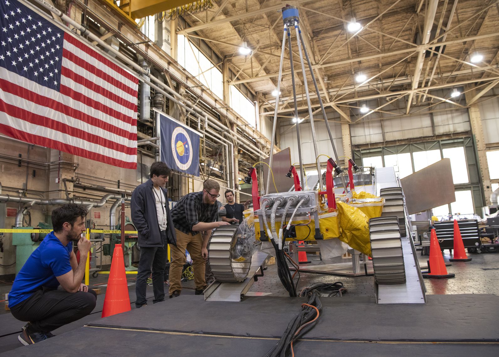 A large room with a model of a rover towards the center and four people examining it to the left. In front of the rover is 