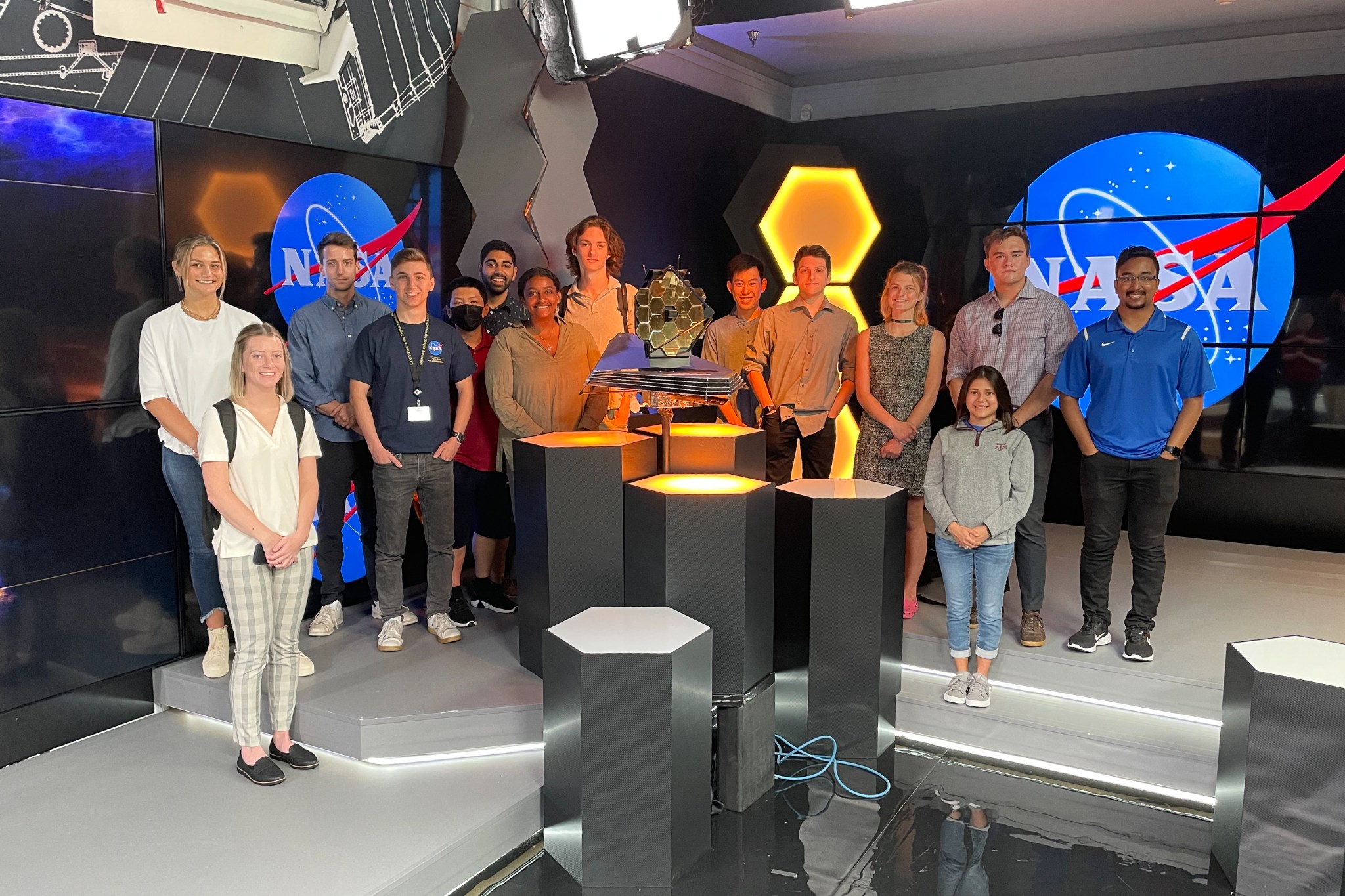 14 young people gather on a stage in front of black glass and NASA logos. In front hexagonal pedastles support a model of the James Webb Space Telescope, with it's golden honeycomb mirror. Hexagons and technical drawings permeate the design.
