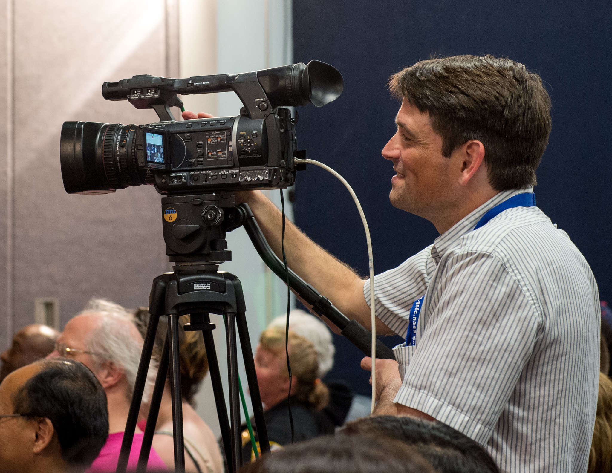 A man smiles while standing behind a video camera.