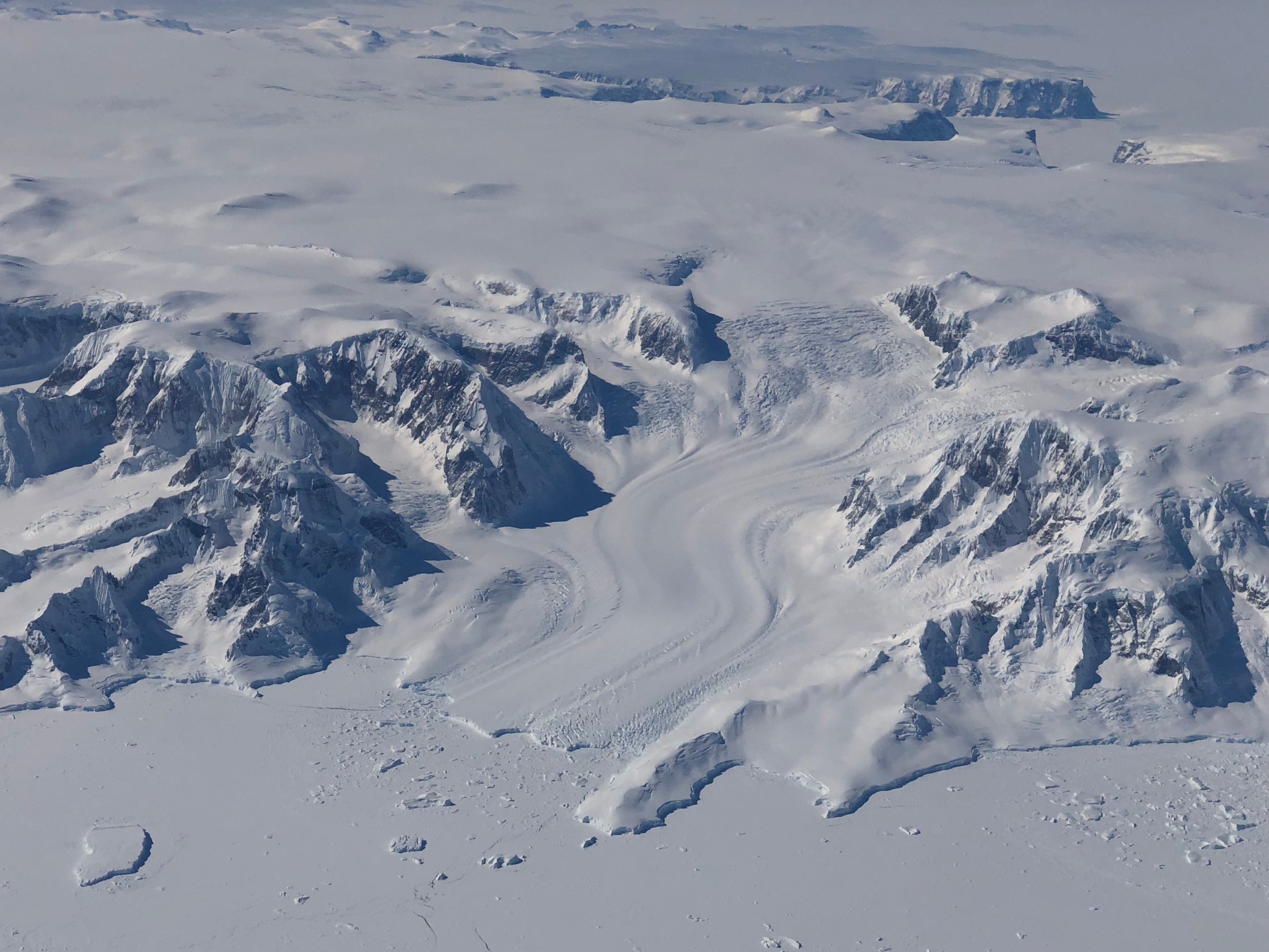 A photo of a glacier on the Antarctic Peninsula, flowing into the Bellingshausen Sea. The glacier looks like a frozen white river winding until it hits the flat extent of the sea ice.