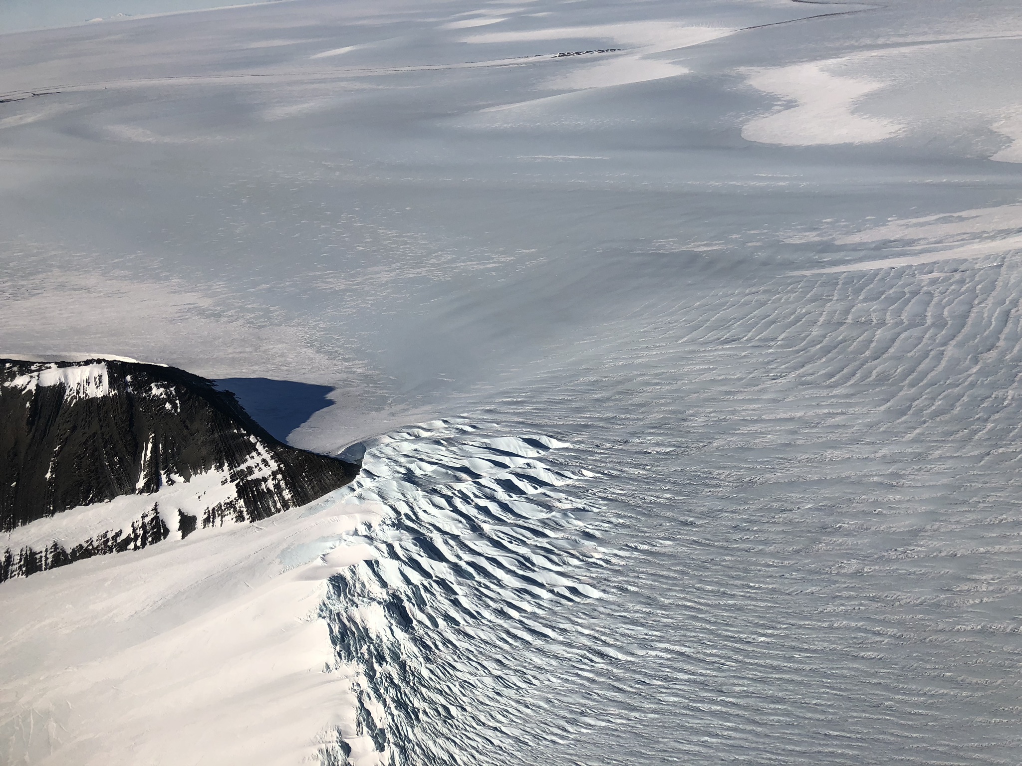 A photograph of crevasses forming as ice flows around an Antarctic mountain. The crevasses look like bumpy, light blue frosting, which meets with smoother, white ice in the left third of the picture.