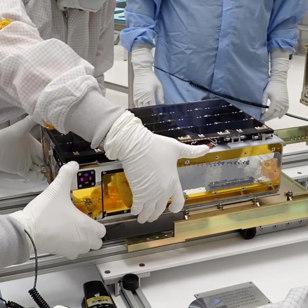 LASP researchers working on CTIM at the University of Colorado, Boulder. About the size of a shoebox, CTIM is the smallest instrument ever dispatched to study total solar irradiance. Gloved hands hold a flat, rectangular instrument with gold along the short side and solar panels along the top.