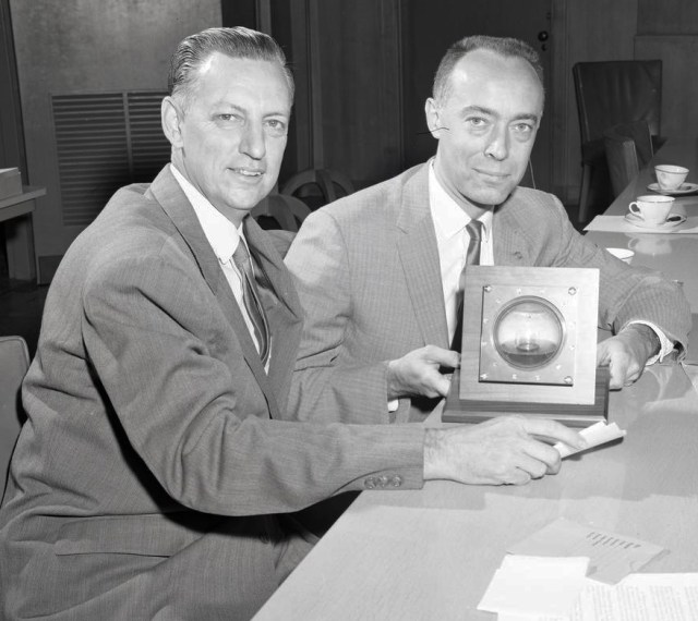 Two men sit at a table holding a microgravity fluids experiment.