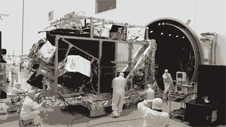 Grayscale, sped-up sequence of technicians in clean room attire moving a satellite into a vacuum chamber and sealing the chamber's large metal door.