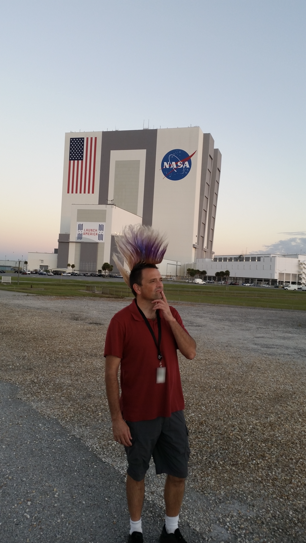 A man wearing a red polo shirt and gray shorts stands looking right. His hair is mostly dark and short except for a purple and orange mohawk. A large building at Kennedy Space Center stands behind him.