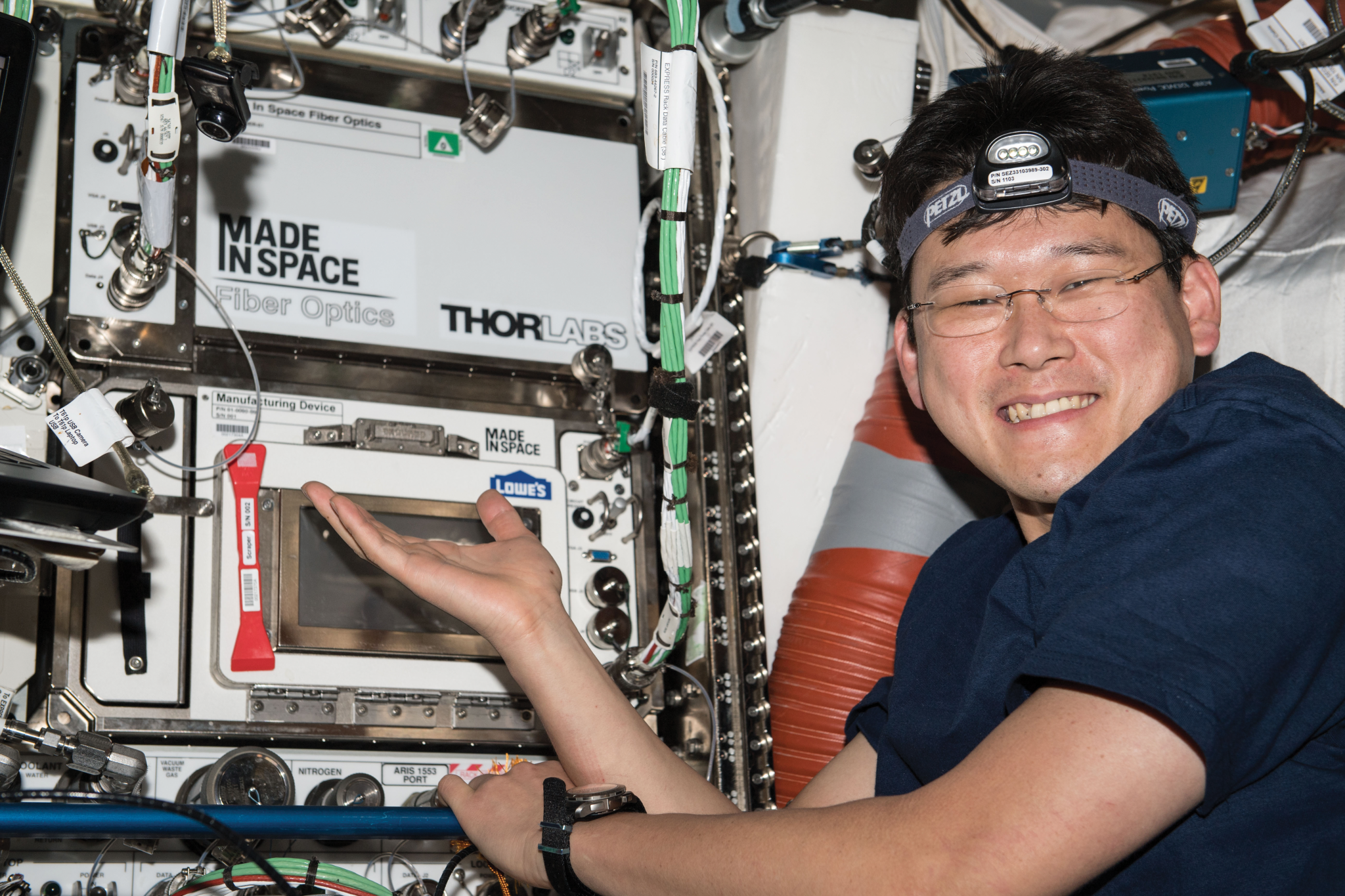 Kanai wears a dark blue short-sleeved t-shirt, a wristwatch, and a headlamp. He is smiling at the camera and gesturing toward hardware that has two microwave oven-sized sections, one above the other. The top one is white and is labeled 