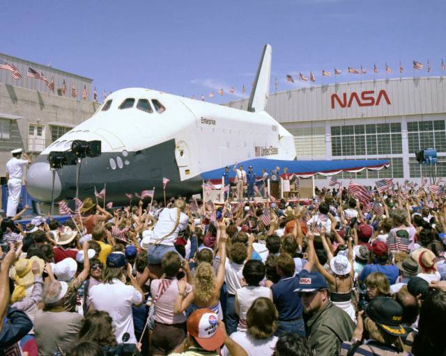 A huge crowd surrounds the Enterprise Shuttle. In the background are buildings, one of which features NASA's worm logo.