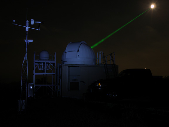 A NASA’s Goddard Space Flight Center Laser Ranging Facility directing a laser (green beam) toward the Lunar Reconnaissance Orbiter (LRO) spacecraft in orbit around the moon (white disk). The moon has been deliberately over-exposed to show the laser.
