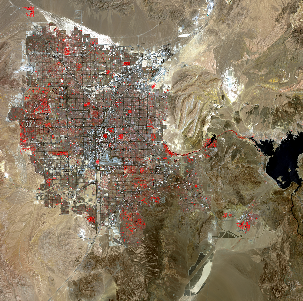 A satellite image of Las Vegas, with the city visible as a sprawling, gridded area of browns and grays with red highlights that denote human-made parks and green spaces, covering the center of the image. Rugged desert landscape is visible as grooved tan and gray-green land surrounding the city, with Lake Mead appearing as a small black body of water at the image's center right.