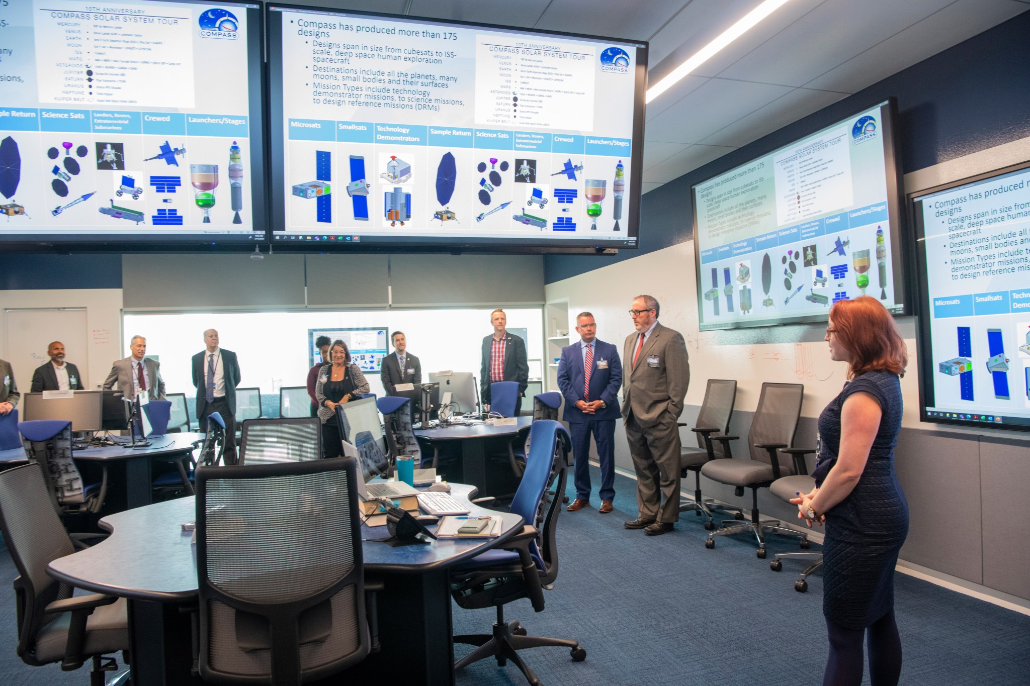 A woman conducts a tour of a room with large monitors mounted high on the walls.