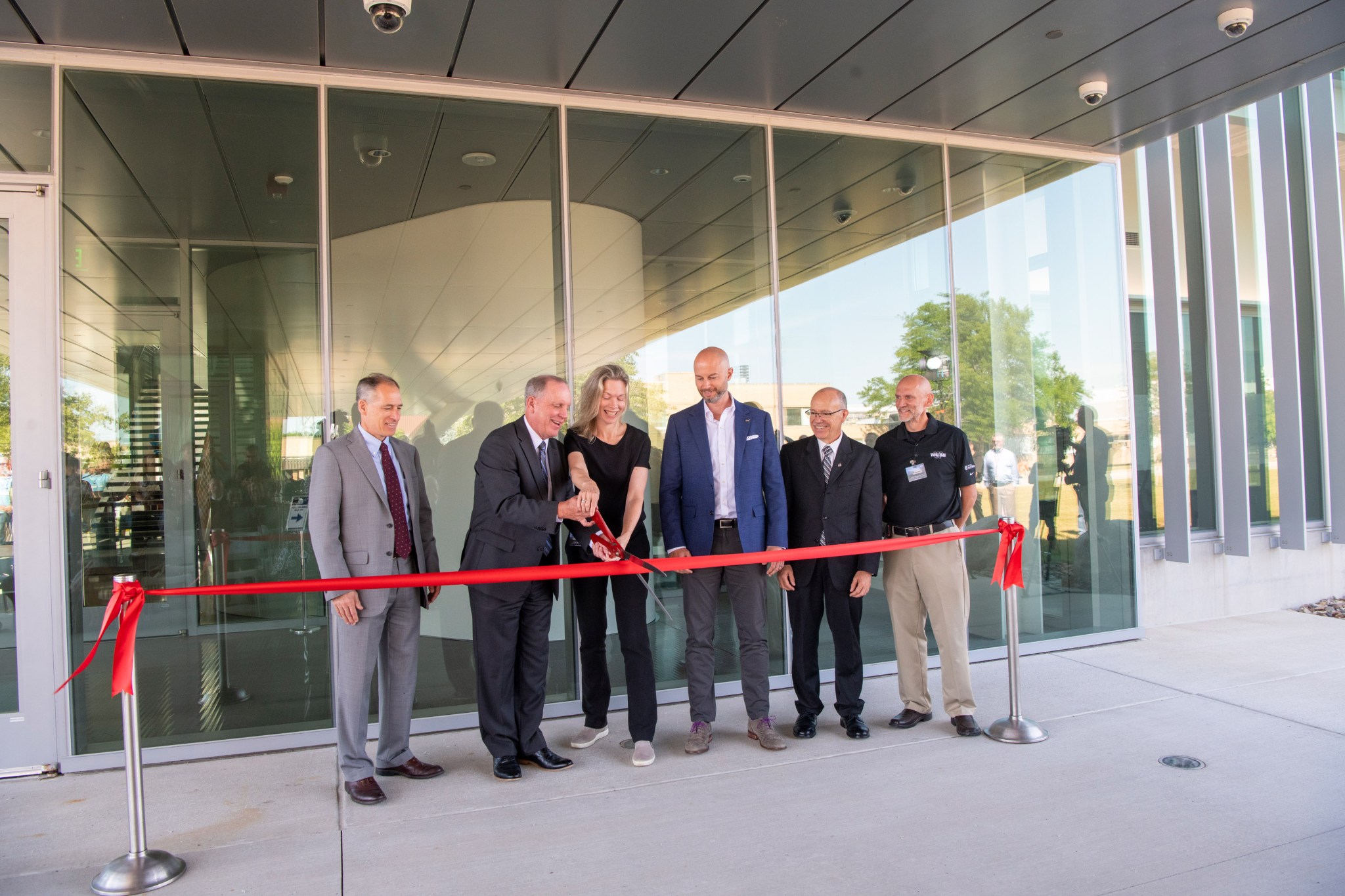 Six people stand behind large red ribbon while two cut the ribbon with large scissors.