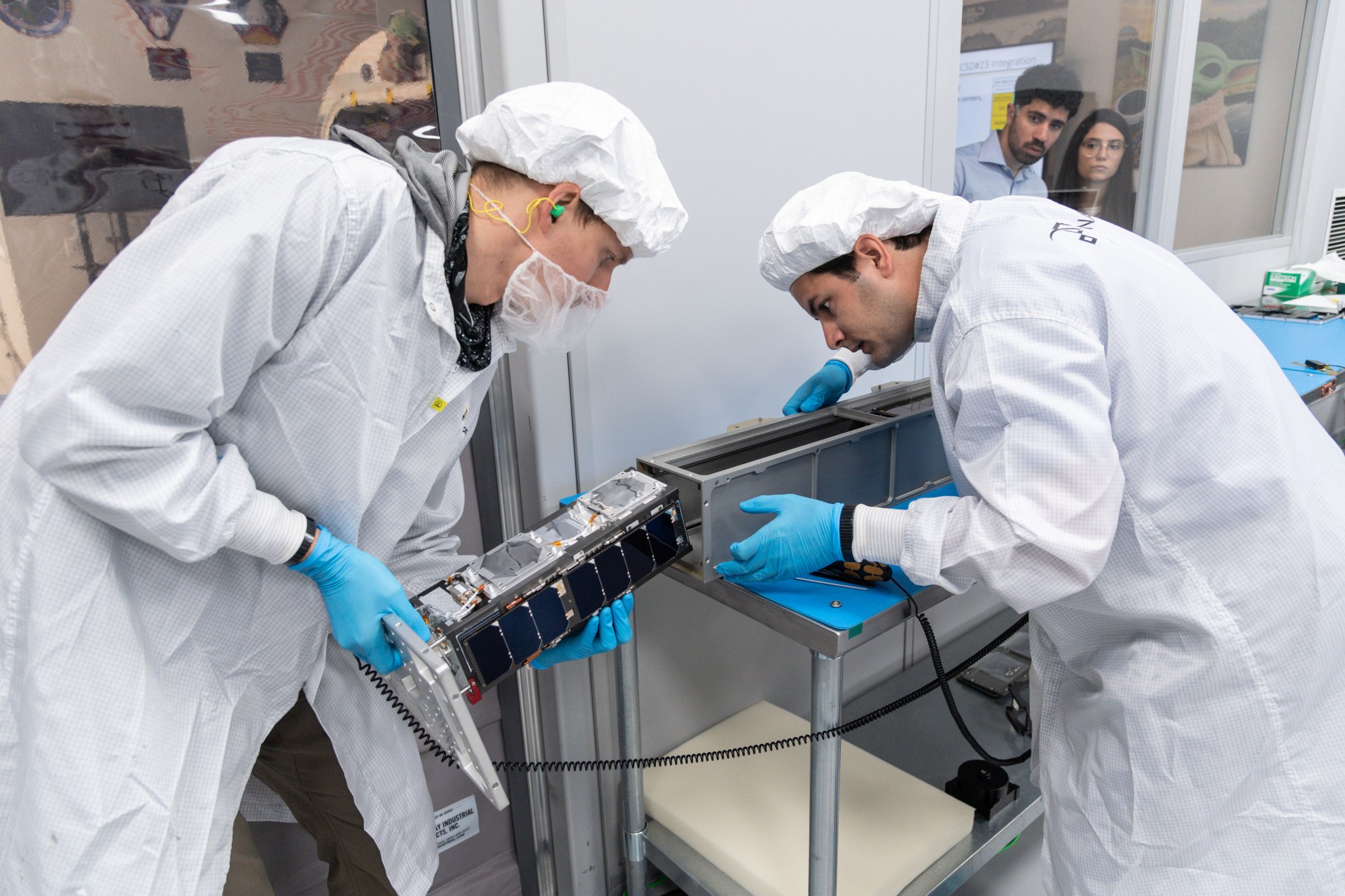 Two technicians wearing lab coats, gloves, and other cleanroom apparel insert a 10 centimeter by 30 centimeter (approximately 4 inch by 12 inch) device into a similarly-sized receptacle while being observed by two people through a window.