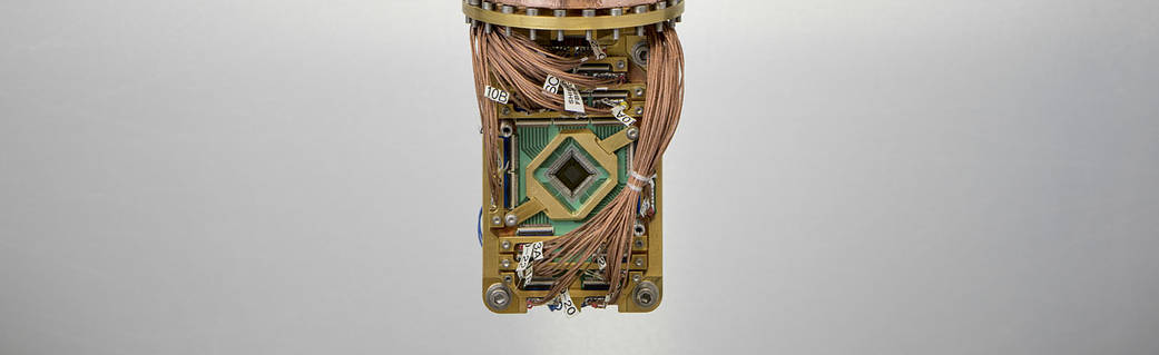 A close-up shot of a computer chip board with a diamond-shaped chip at the center, framed in gold metal, with bundles of copper wiring attached at the top and bottom.