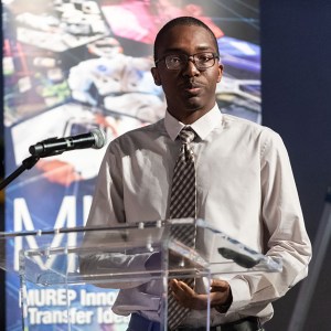 A MITTIC participant stands at a podium and presents to a panel of judges with the quote