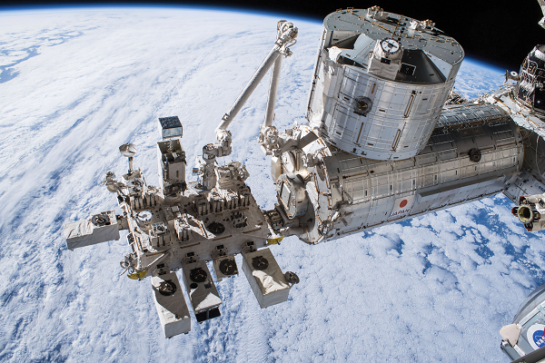 The cylindrical Kibo module to the right of this image has another cylindrical module on its top and a collection of instruments attached to a platform extending out to the left. Four large white boxy shapes extend out to the front and others to the back, including MAXI, which is at the end of the Japanese robotic arm attached to the end of KIBO, visible at the top. The instruments float above a cloudy Earth.