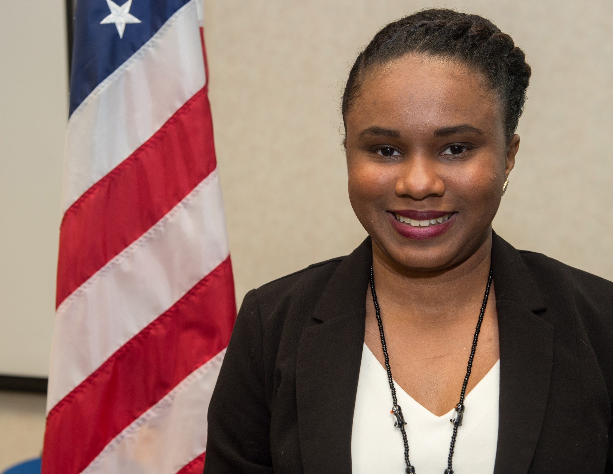 A woman with brown skin and braided black hair smiles at the camera, next to an American flag.