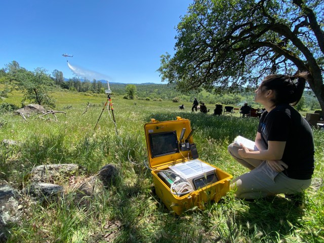 A person kneels in the grass next to a yellow case containing hardware, while a helicopter drops water in the distance