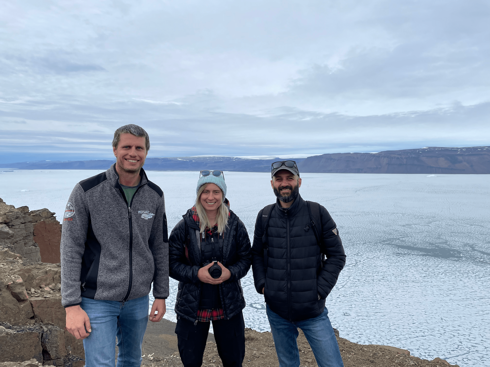 NASA ice scientists Nathan Kurtz, left, Rachel Tilling and Marco Bagnardi stand on a coast of rocky dirt. Behind them sea ice stretches out into the distance. 