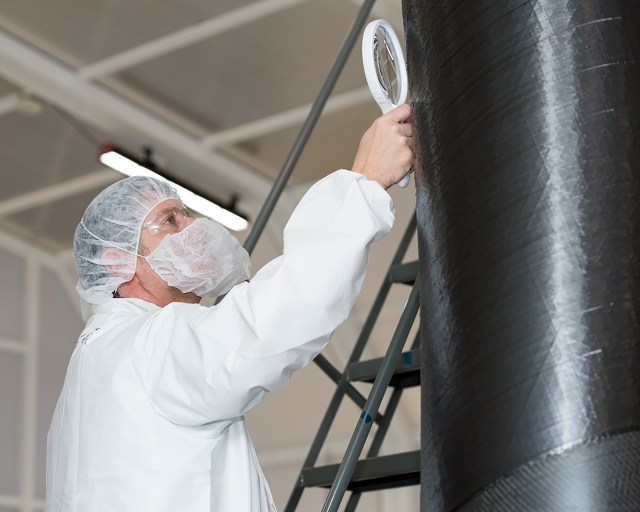 A researcher wearing clean room gear, uses a magnifying glass to check a composite structure.