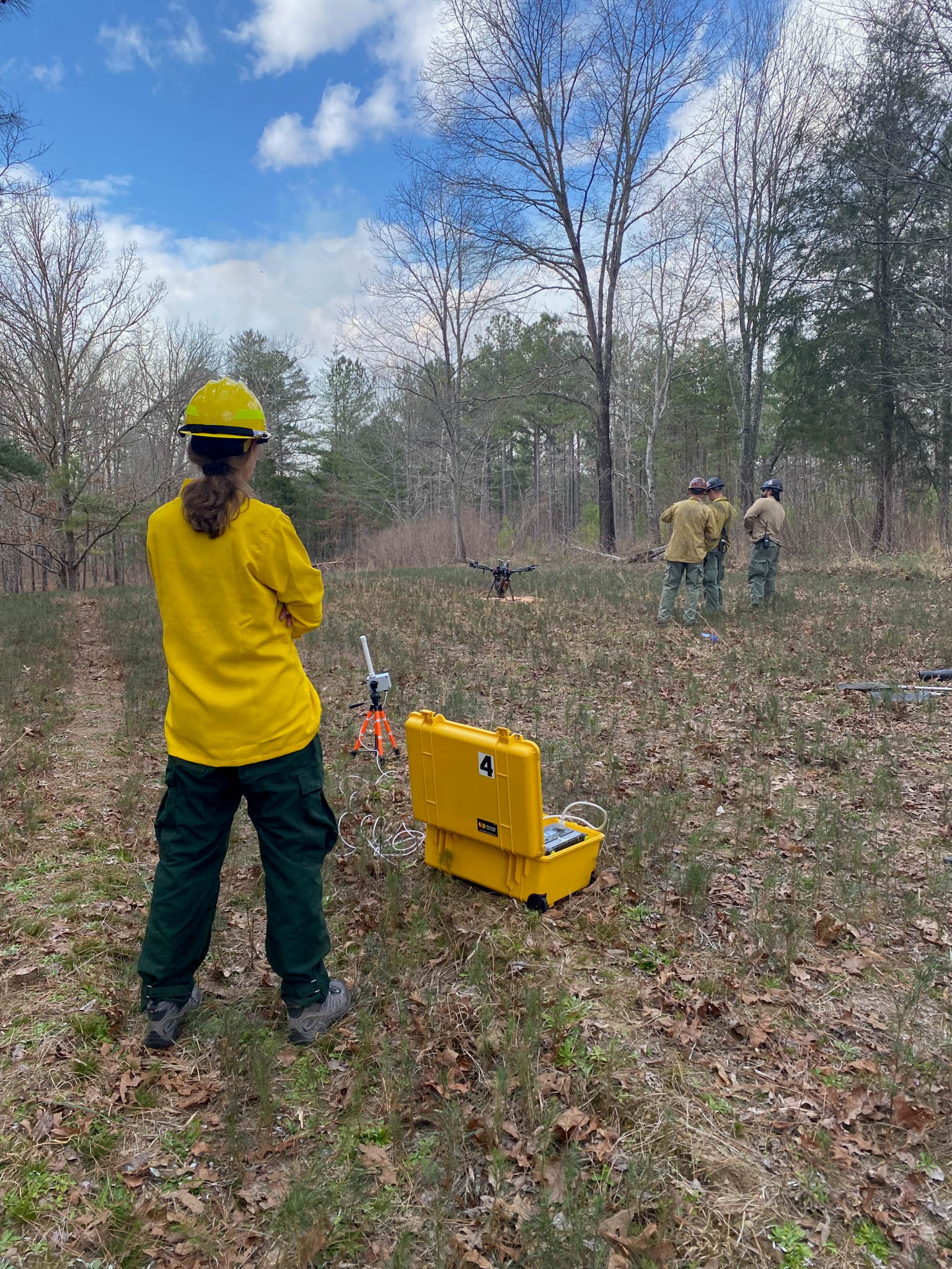 A person in a yellow hard hat stands near a yellow case on the ground, while three people in hard hats observe a drone in the background