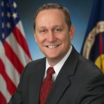 Portrait of Steve Rader wearing a black suit and red tie in front of a blue background. The American flag is to his right and the NASA flag is to his left.