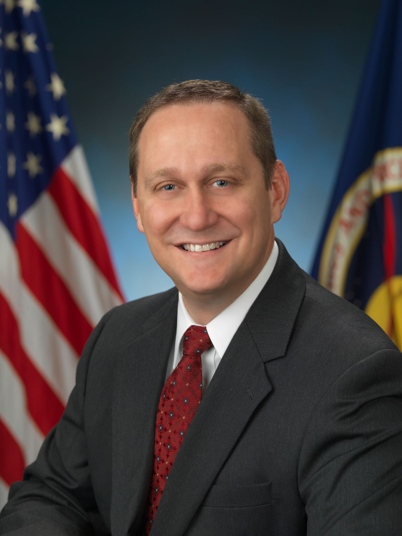 Portrait of Steve Rader wearing a black suit and red tie in front of a blue background. The American flag is to his right and the NASA flag is to his left.