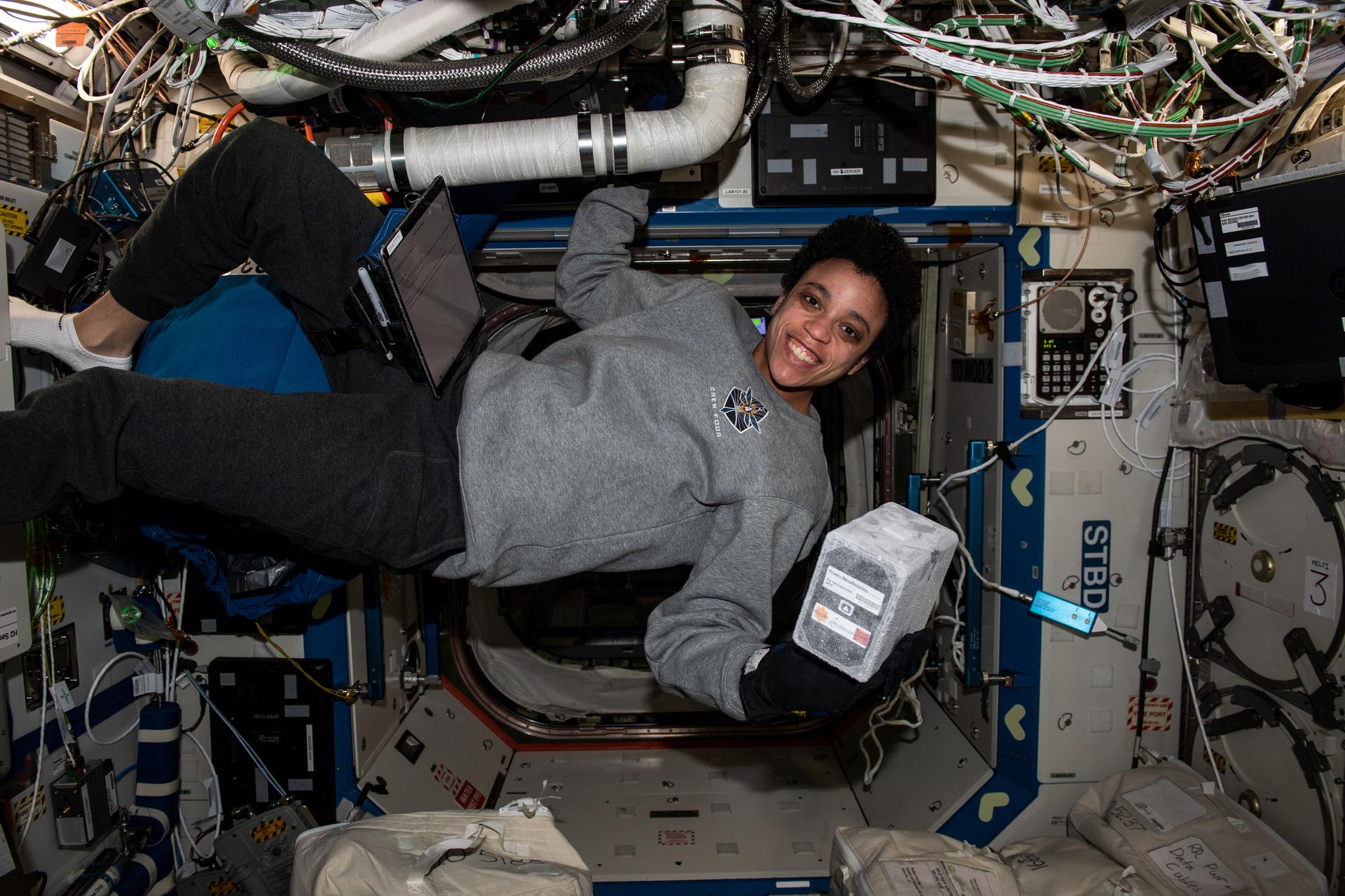 Watkins, smiling at the camera, wears a gray sweatshirt, black pants, and black gloves. She floats in space, holding on to a wall with her right hand and gripping an ice-covered black box about the size of a box of tissues. There is a laptop Velcroed to her leg and a lot of cables over her head.
