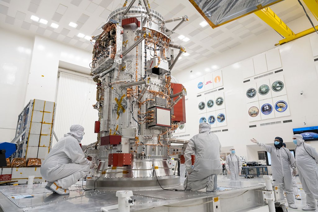 The core of NASA?s Europa Clipper stands in High Bay 1, the largest clean room at the Jet Propulsion Laboratory. Engineers and technicians in white clean room suits stand around it. Some of them kneel while they work and others stand, looking or pointing.