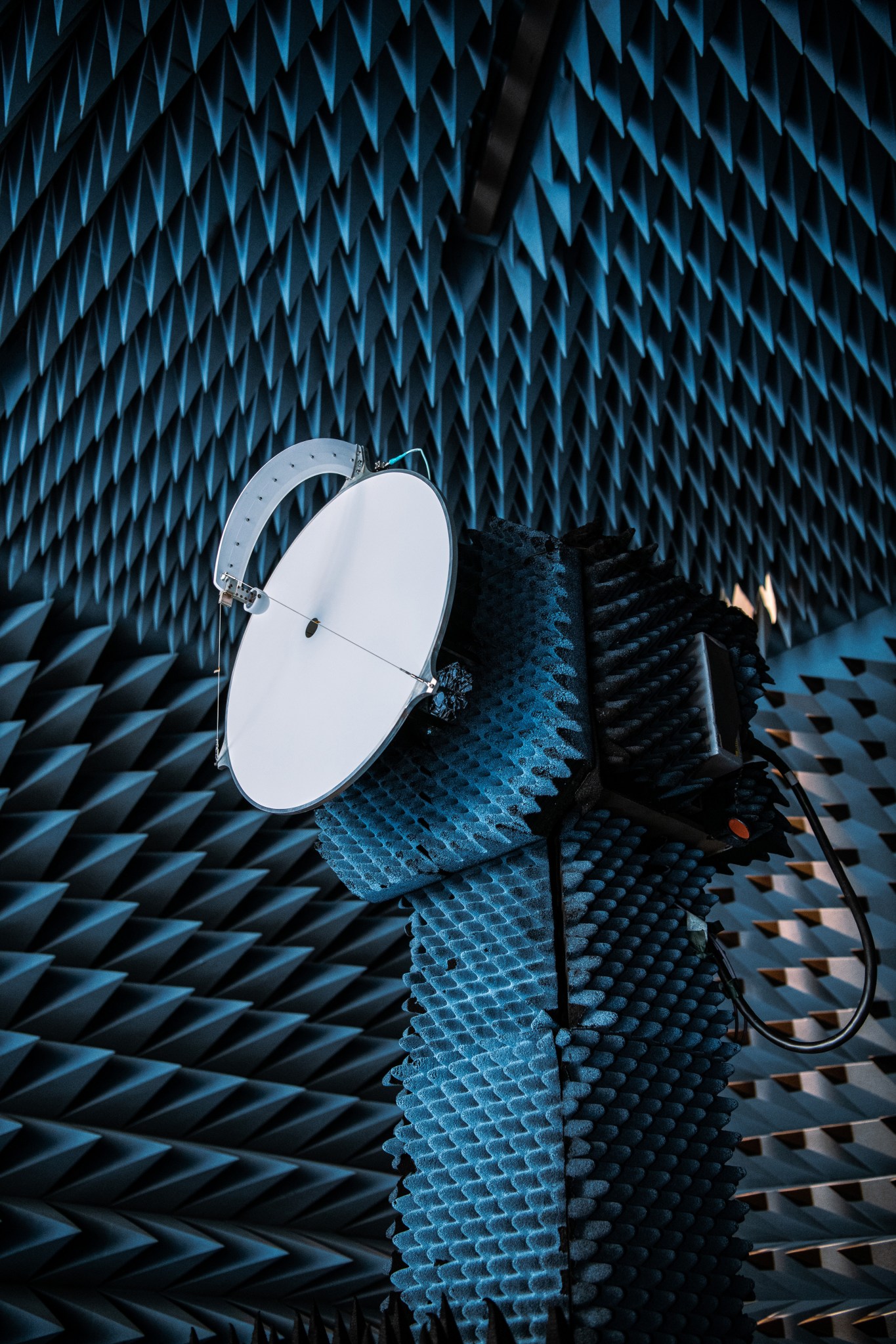 Testing of the PACE Earth Coverage Antenna. A circular white disk with a curved strip extending from its center to the edge of the disk is mounted on a stand, covered with gray foamy spikes to absorb sound. The entire room is also covered in gray foamy spikes, including the ceiling.