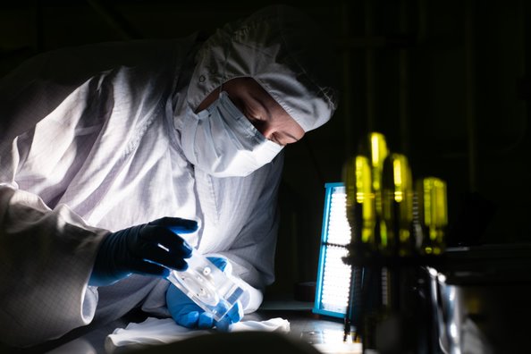 Physical Science Technician Kristen Washington performs a contamination inspection of the OCI Flight Fold Flat Mirror optic. She is wearing a white clean room u0022bunny suit,u0022 including a white hood over her hair, a white mask over the lower half of her face, and blue gloves. It's dark in the room, and she is holding a white piece of technology in both hands, tilted toward a bright light source. In the foreground, a number of screwdrivers with transparent yellow handles are sitting tip-down.