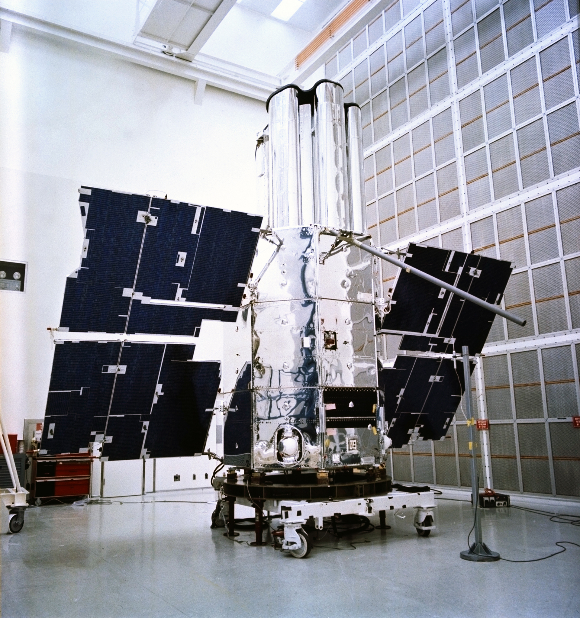 Blue-black solar panels extend from the spacecraft's metal body, which reflects the brightly lit interior of its clean room.   