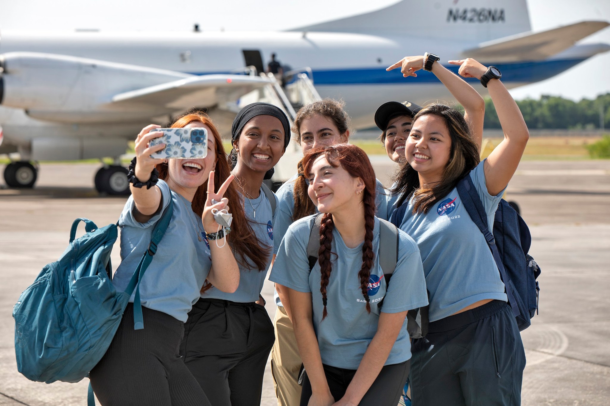 Six college students wearing light blue t-shirts with NASA logo pose taking a selfie in front of NASA's P-3 research aircraft