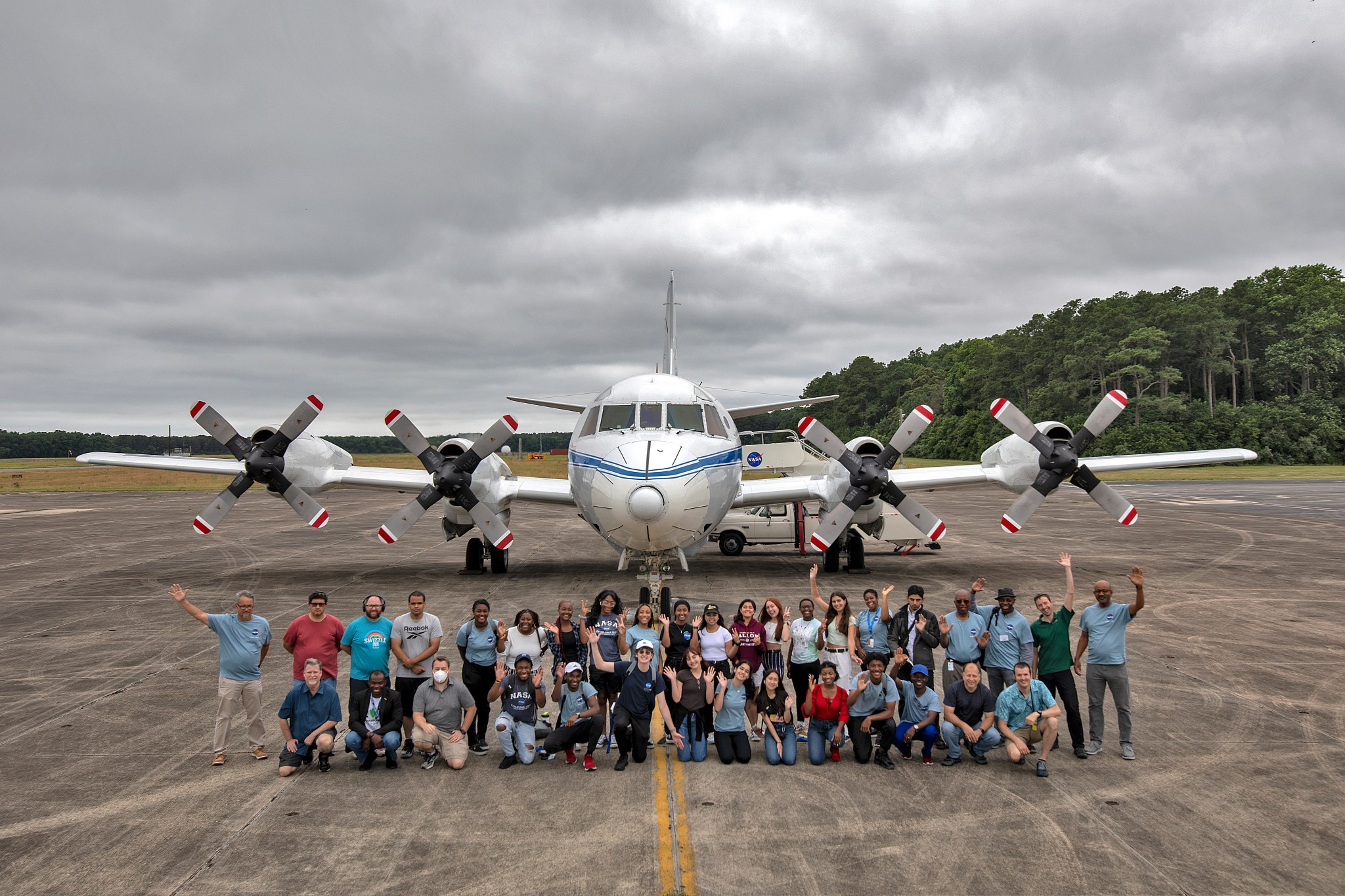 A group of people pose in front of an aircraft facing forwards with four propellers along its wings