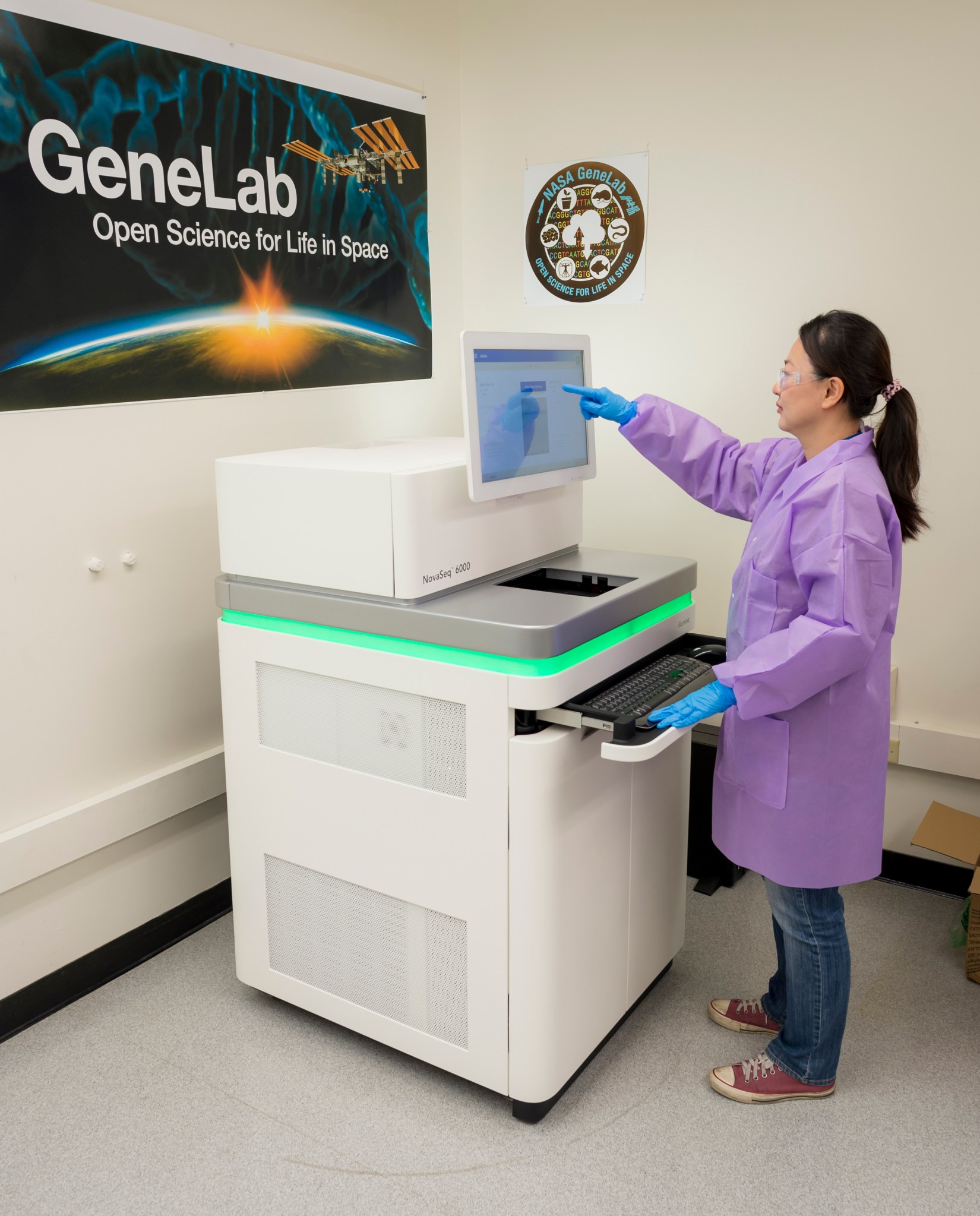 A woman stands at a machine and points to a monitor, a banner for GeneLab is on the wall in the background.