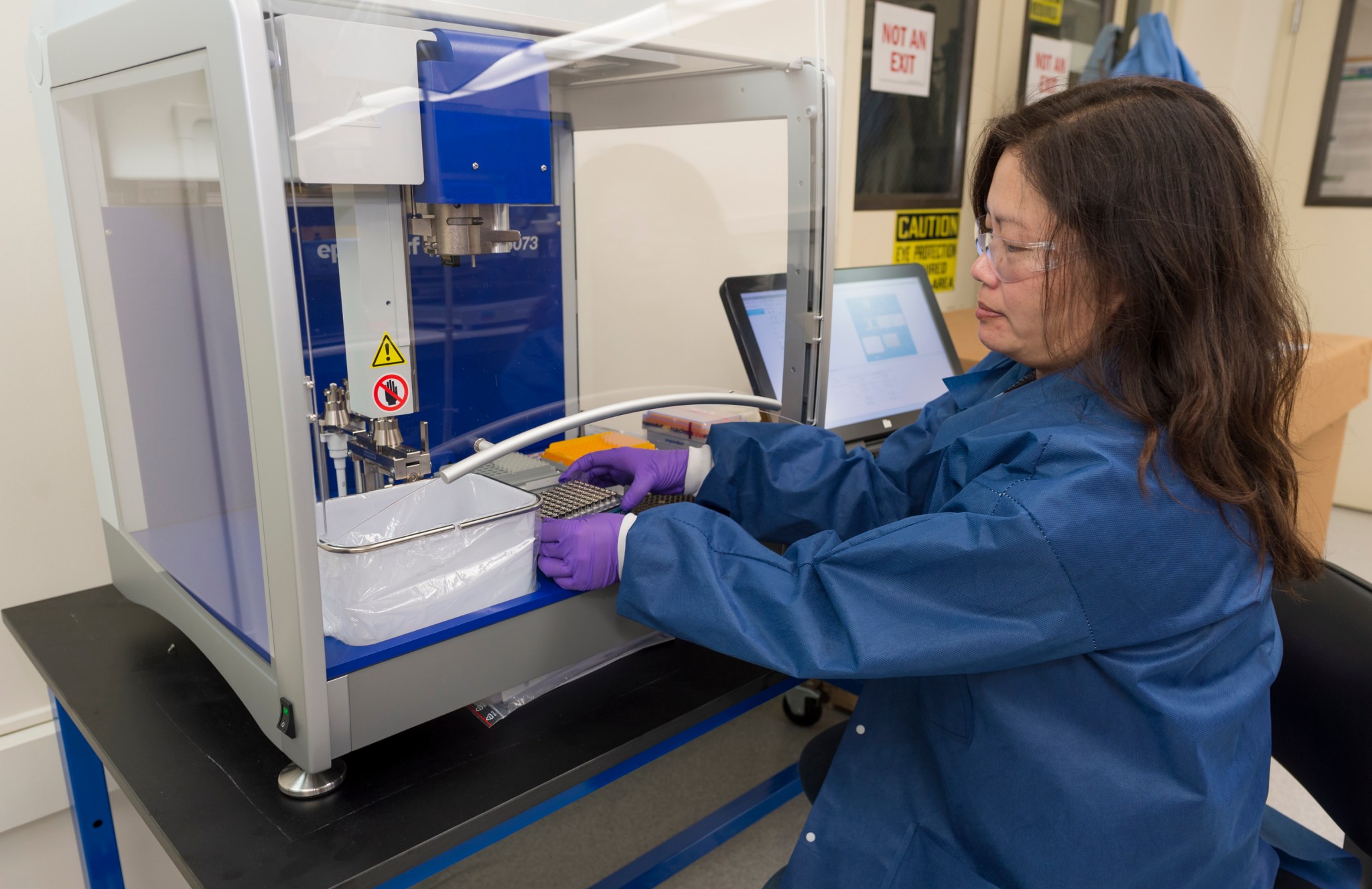 A person wearing a dark blue lab coat and purple gloves works with a plate of biological samples 