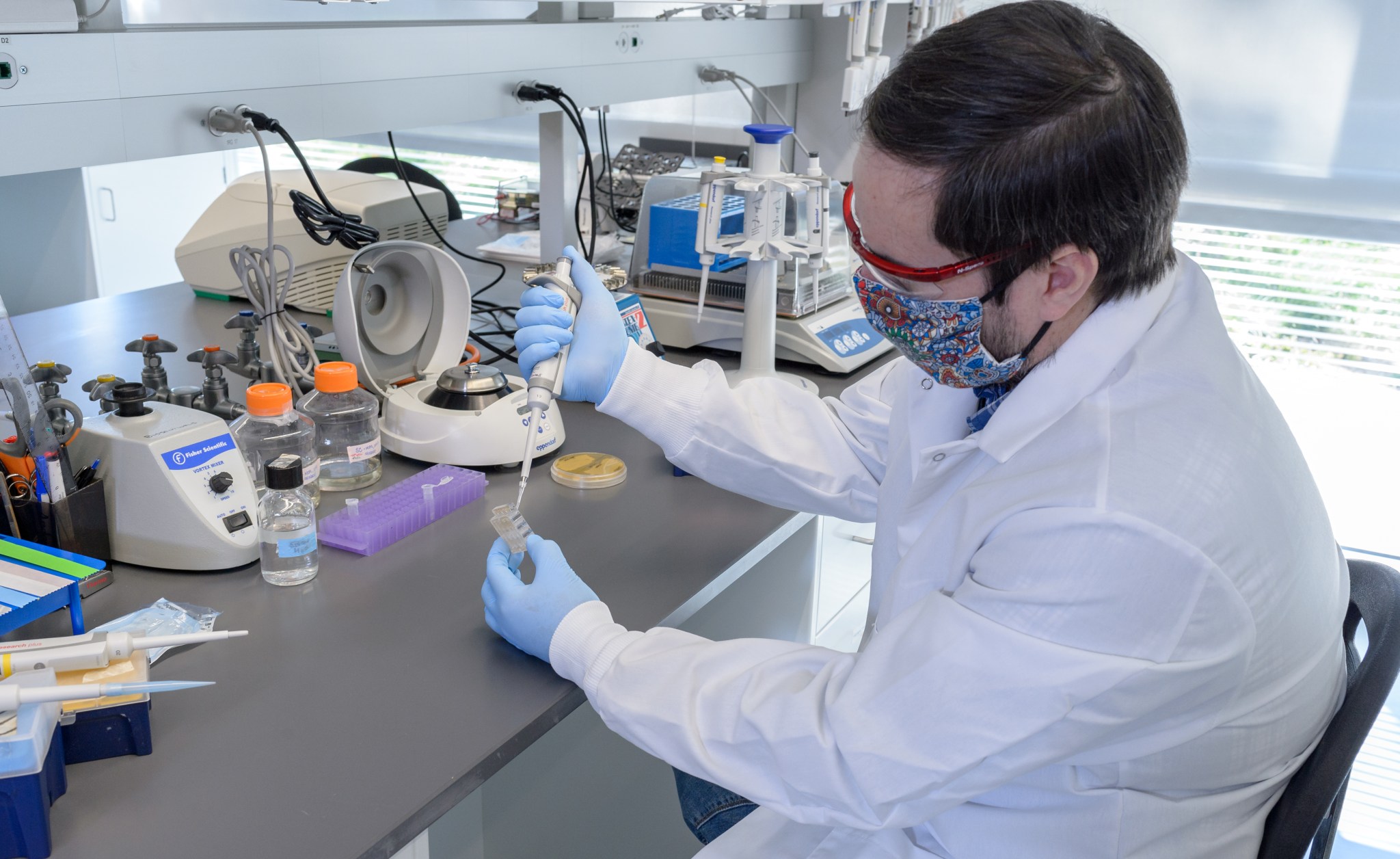 A person in a white lab coat and gloves loads a fluidic card with yeast, similar to the cards used in the BioSentinel science payload.n