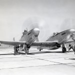 Aircraft with two pilots inside, propellers in motion, watch as ground crew guide them on taxiway.