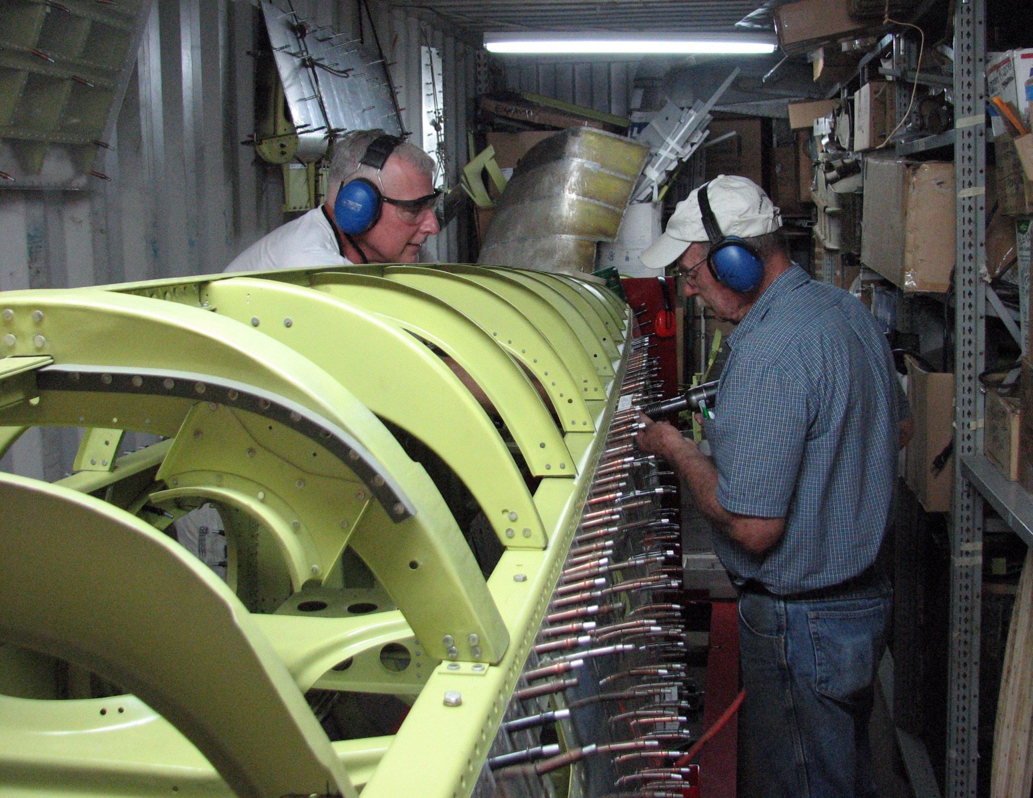 Two men, outfitted with headphones, face each other as they work on rivets to an aircraft fuselage.