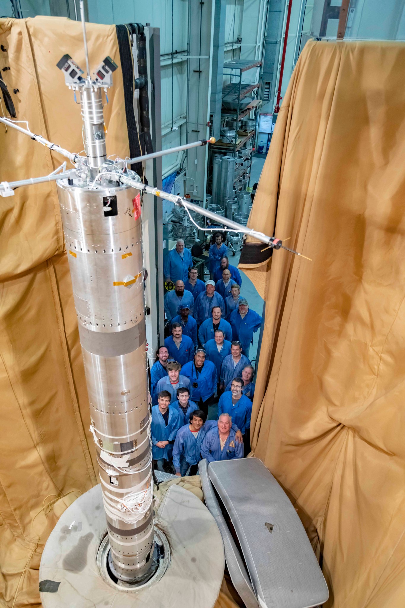 In the foreground, a section of a rocket payload stands vertical on a platform, surrounded by brown cloth. Below the rocket in the background is a line of people in blue jackets looking up at the camera for a group photo.